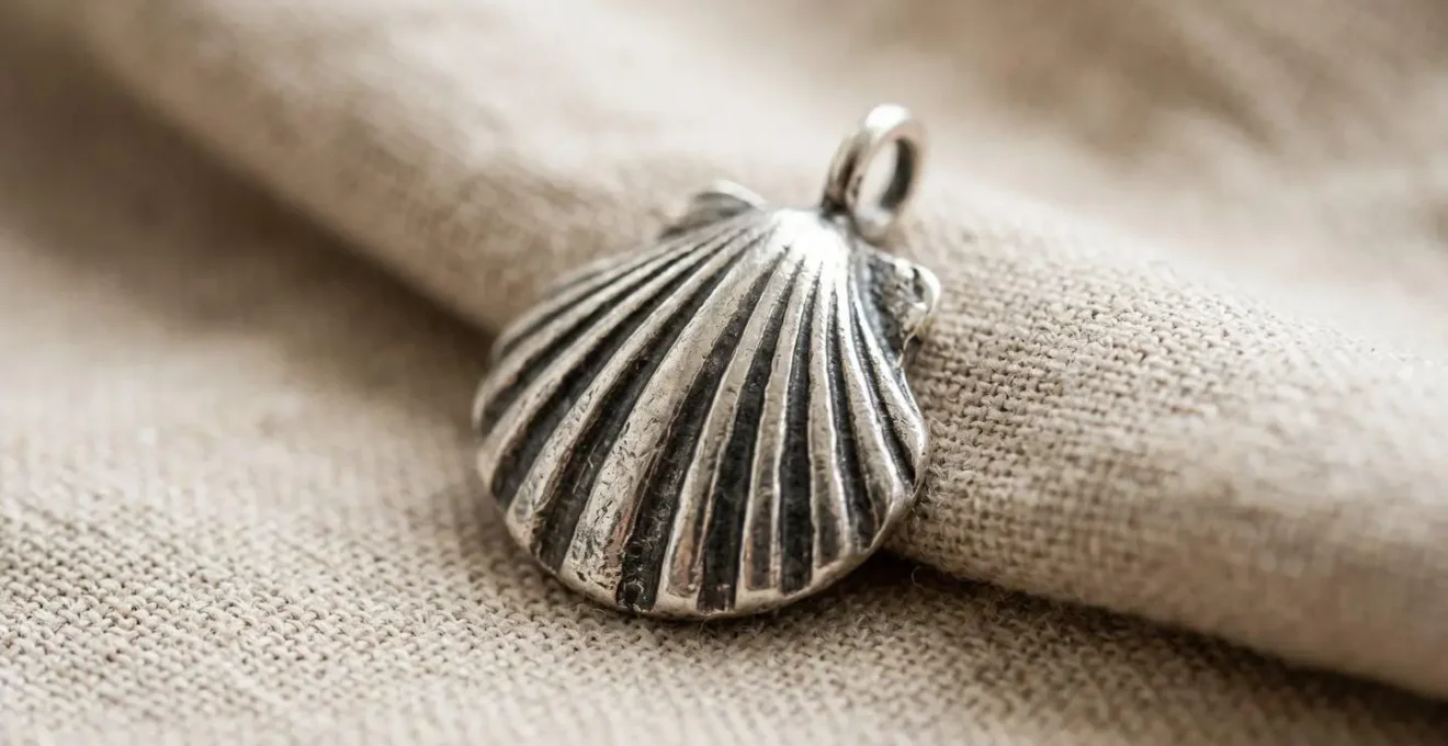 Detalle macro de charm de concha de plata representando el Camino de Santiago con textura realista sobre superficie suave